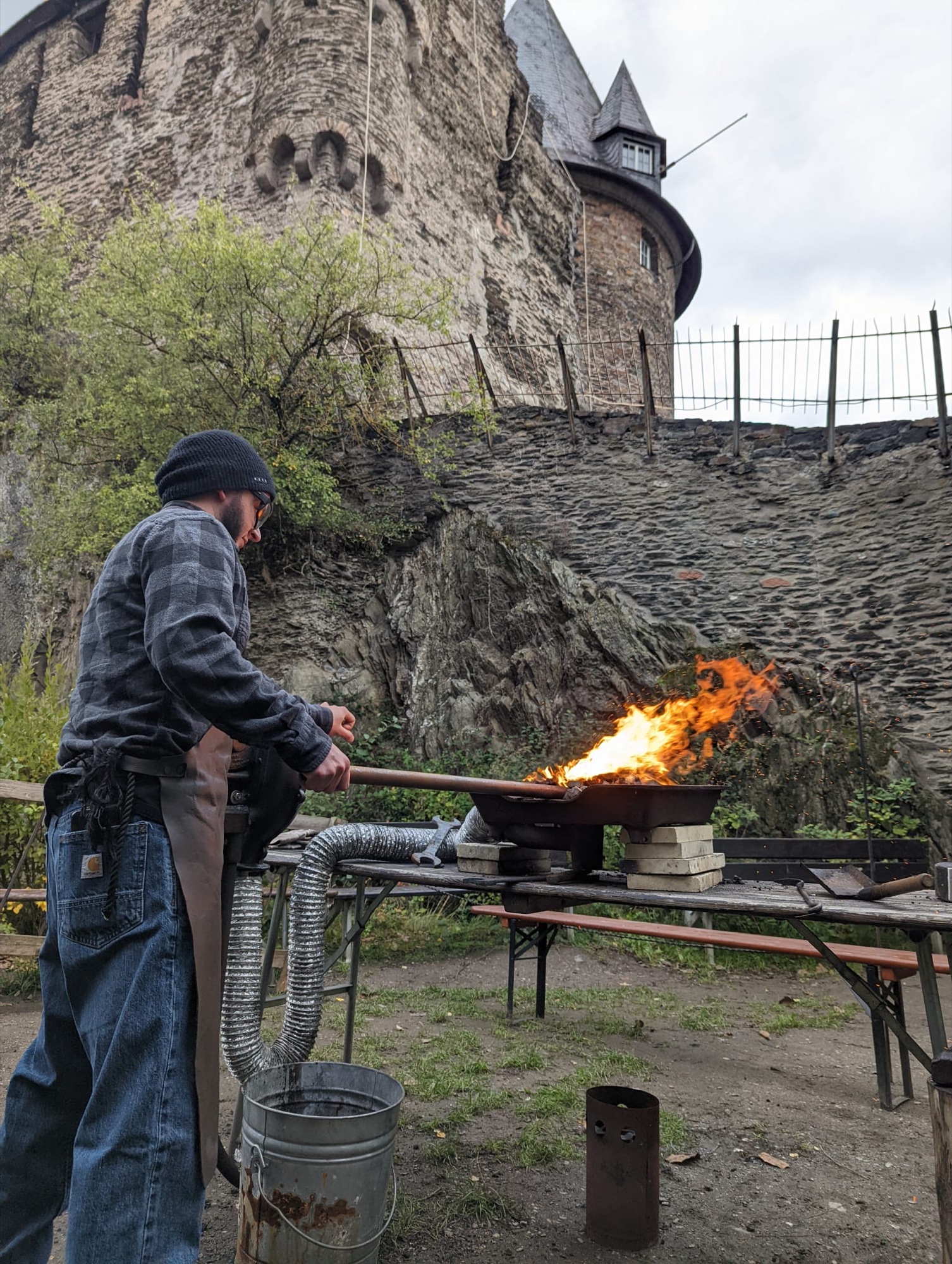 A blacksmith working a bright outdoor fire beside a German castle